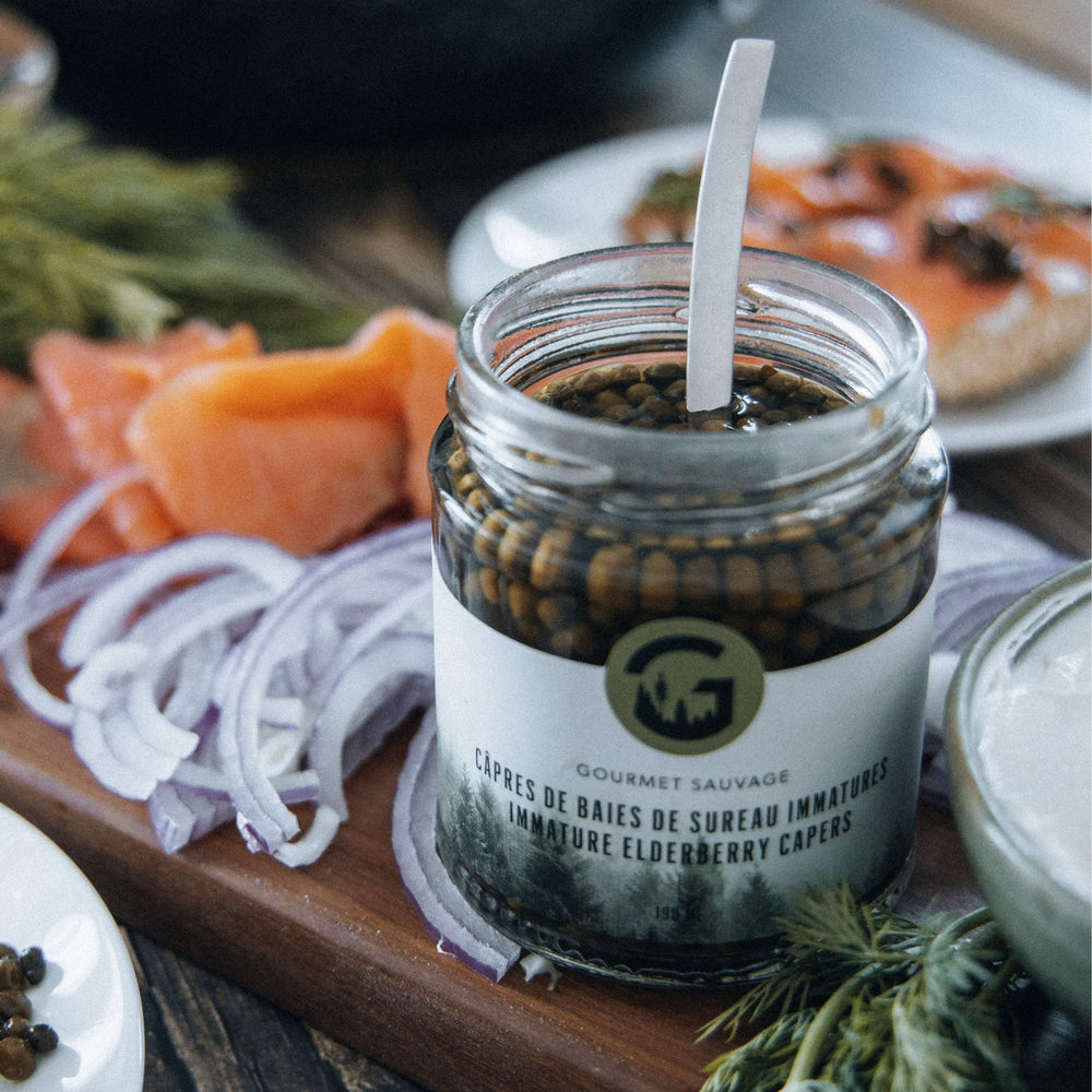 
                      
                        open jar of elderberry capers seen in situ on a wooden board alongside sliced red onion, smoked salmon, and dill
                      
                    