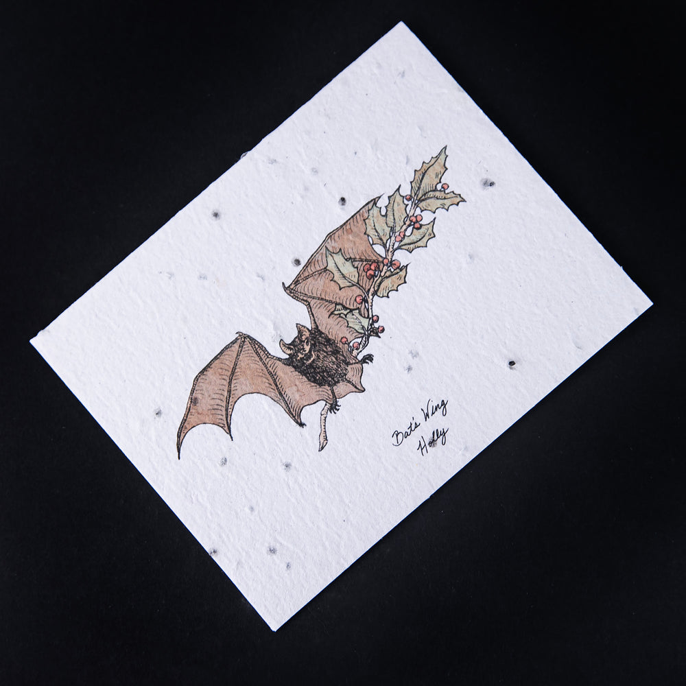 Cream-coloured card with illustration of a bat holding a branch of holly. The paper is naturally textured and studded with seeds.