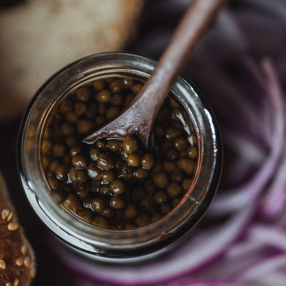 
                      
                        close up of open jar of elderberry capers with a wooden spoon in it
                      
                    