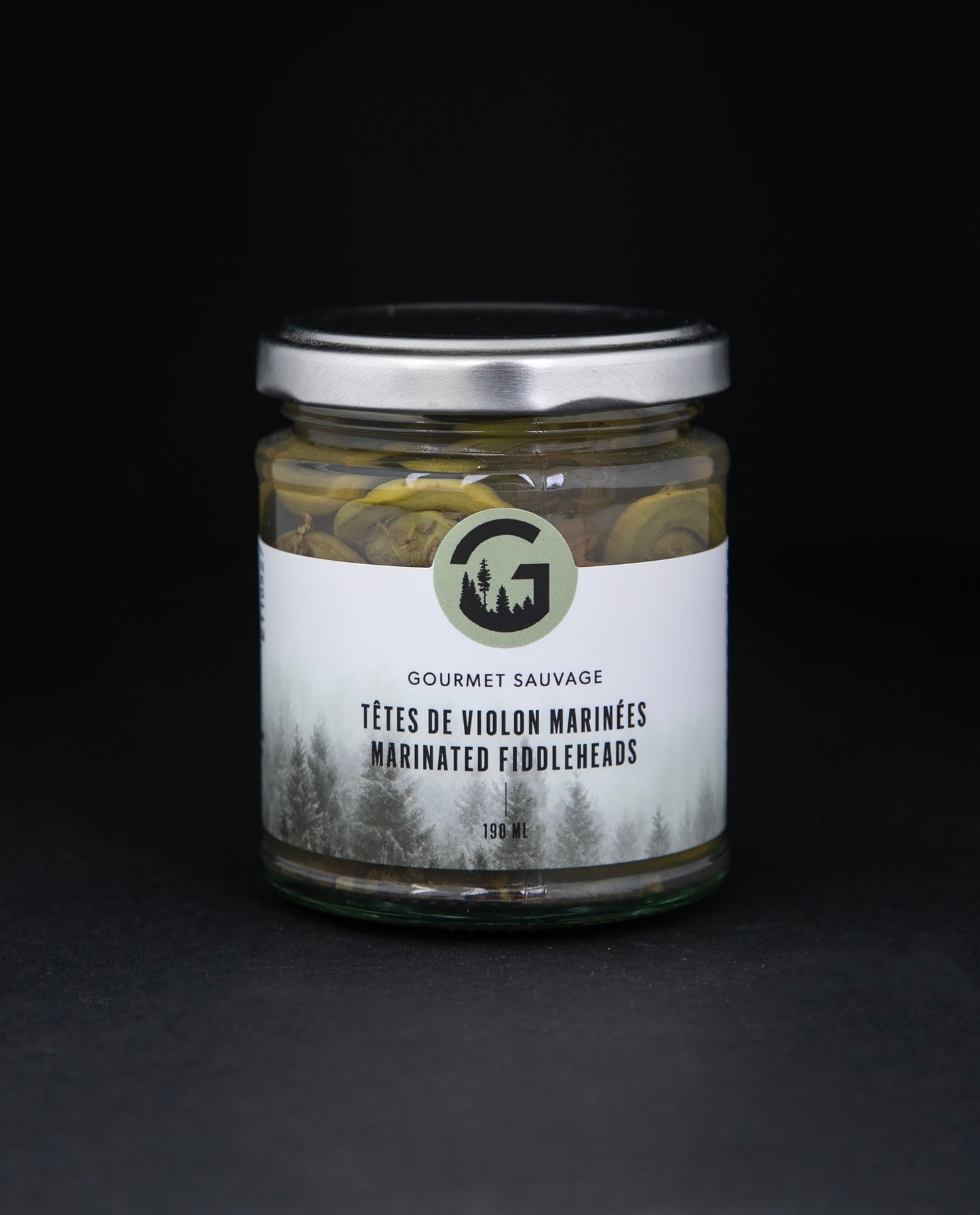 Clear glass jar with metal lid of Gourmet Sauvage's marinated fiddleheads, sitting on black background