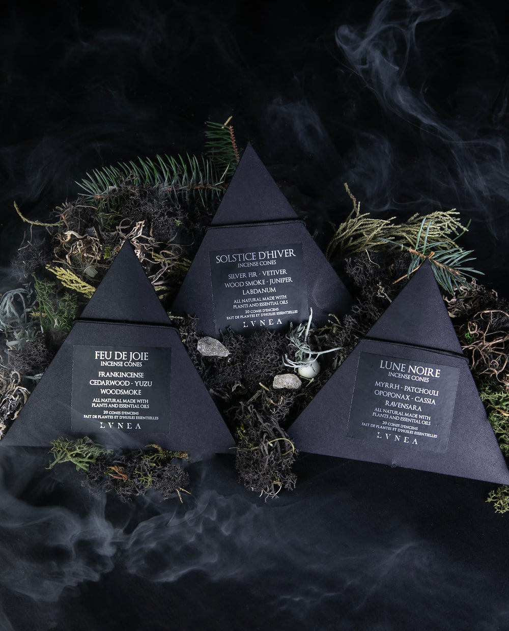 A set of 3 boxes of LVNEA incense cones, sitting in a bed of moss and evergreens, with wisps smoke curling around them. The boxes are pyramid-shaped and made of black cardboard.