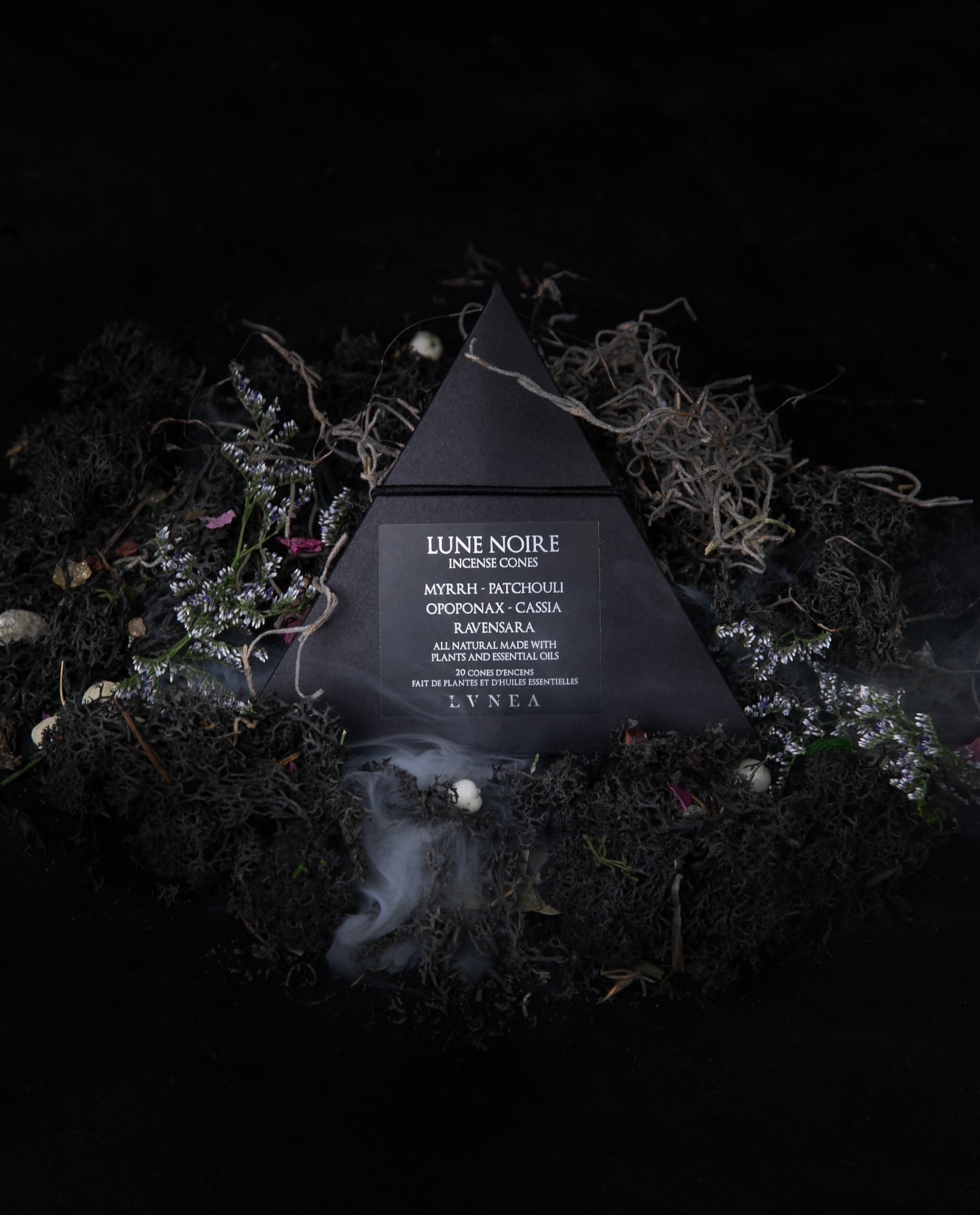 Closed box of LVNEA's 'Lune Noire" incense sitting on a bed of moss and dried florals on a black background, with a wisp of smoke in the foregroud