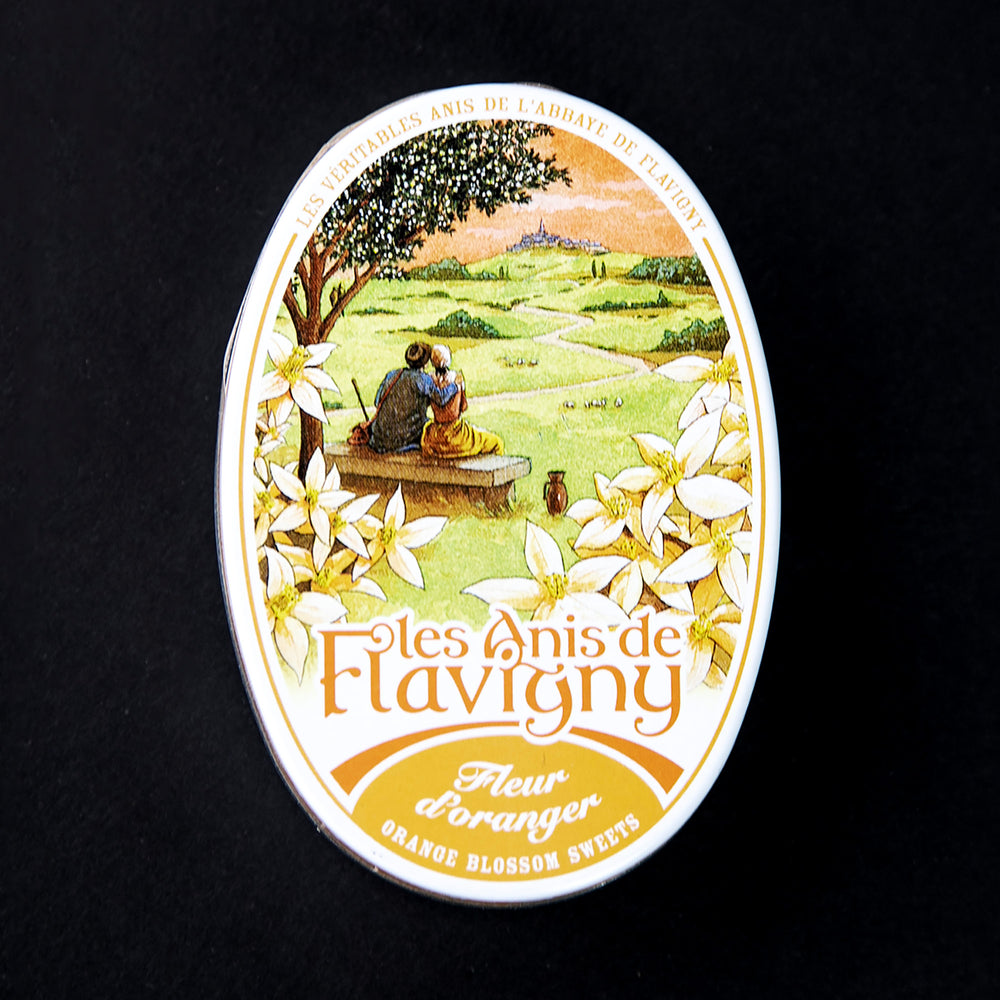 Closed tin of Anis de Flavigny bonbons. The lid is ornate and features illustrations of orange blossom flowers and a pastoral scene of two people sitting on a bench and gazing out at the countryside.