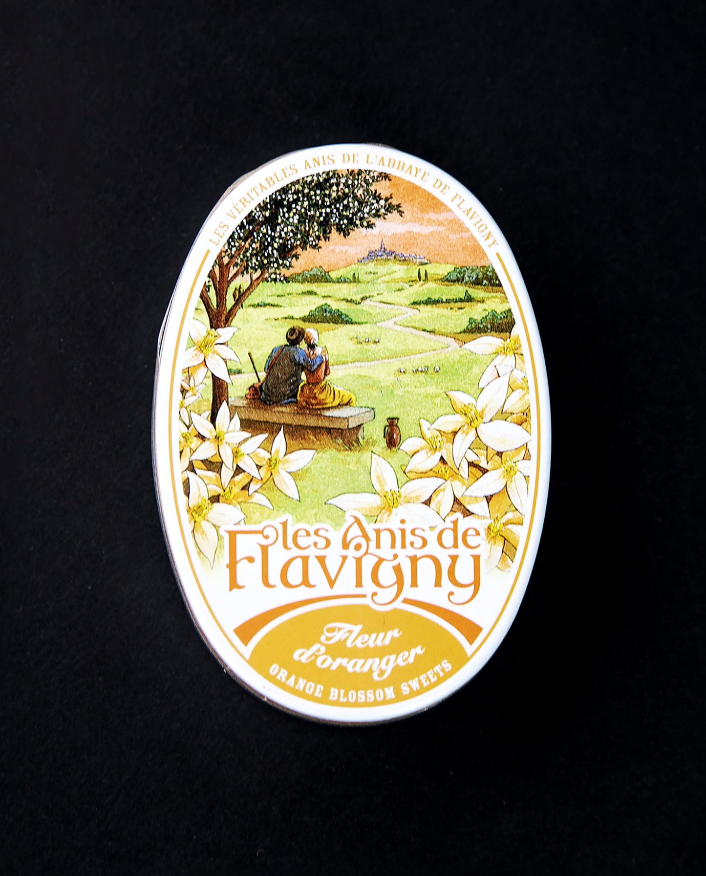 Closed tin of Anis de Flavigny bonbons. The lid is ornate and features illustrations of orange blossom flowers and a pastoral scene of two people sitting on a bench and gazing out at the countryside.