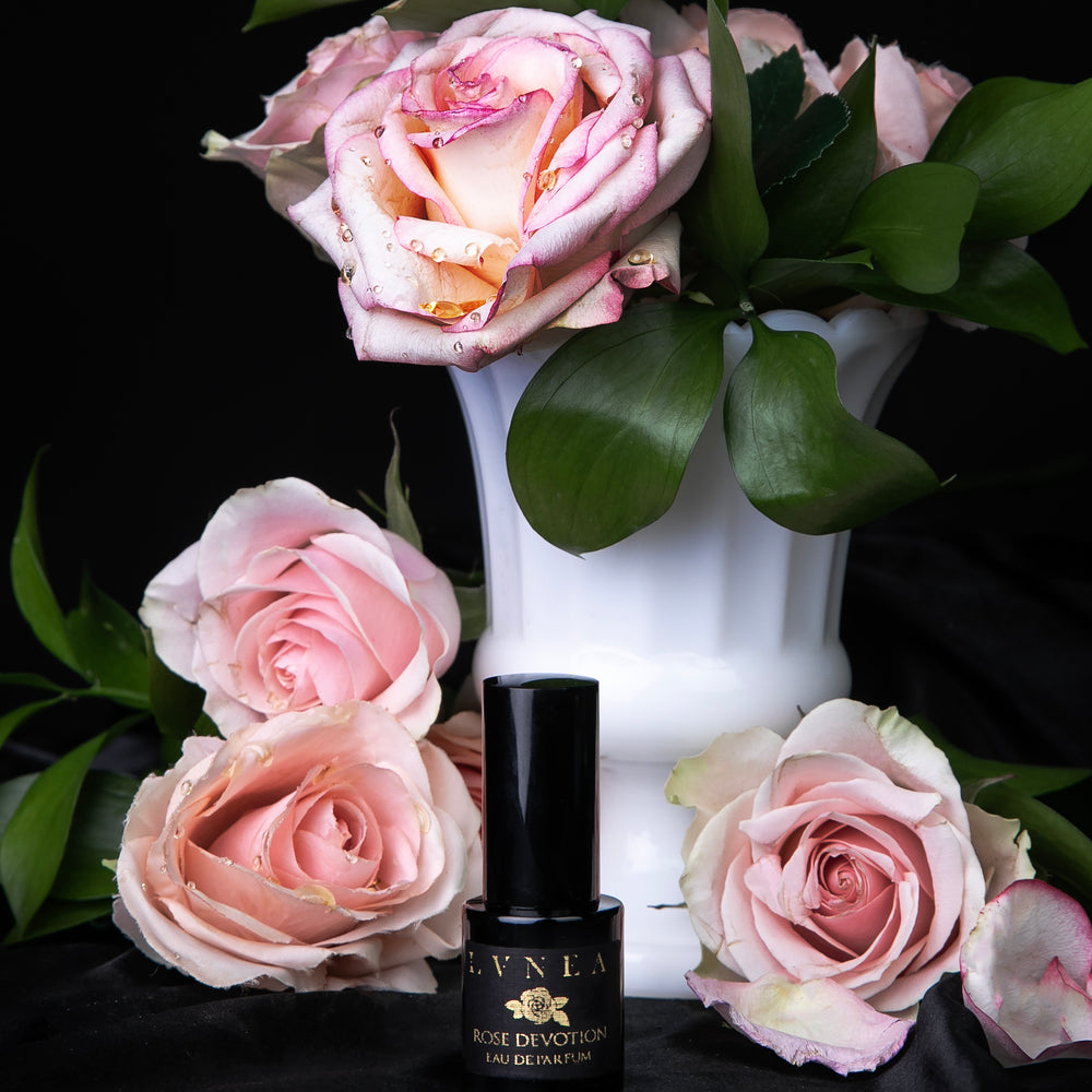 15ml black glass bottle of LVNEA's "Rose Devotion" limited edition eau de parfum, against black satin and surrounded by blush-coloured roses and greenery