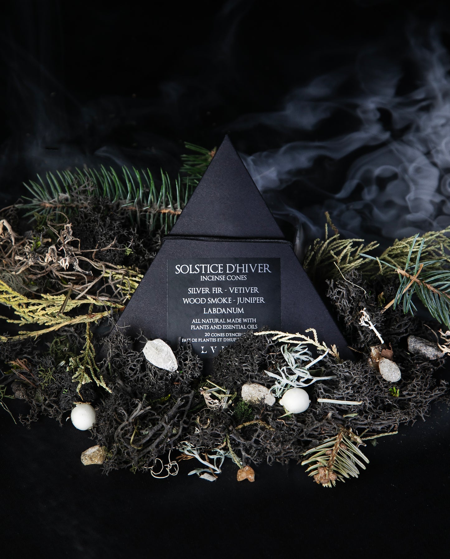 Closed box of LVNEA's 'Solstice d'Hiver" incense sitting on a bed of moss and conifers on a black background, with a wisp of smoke in the background