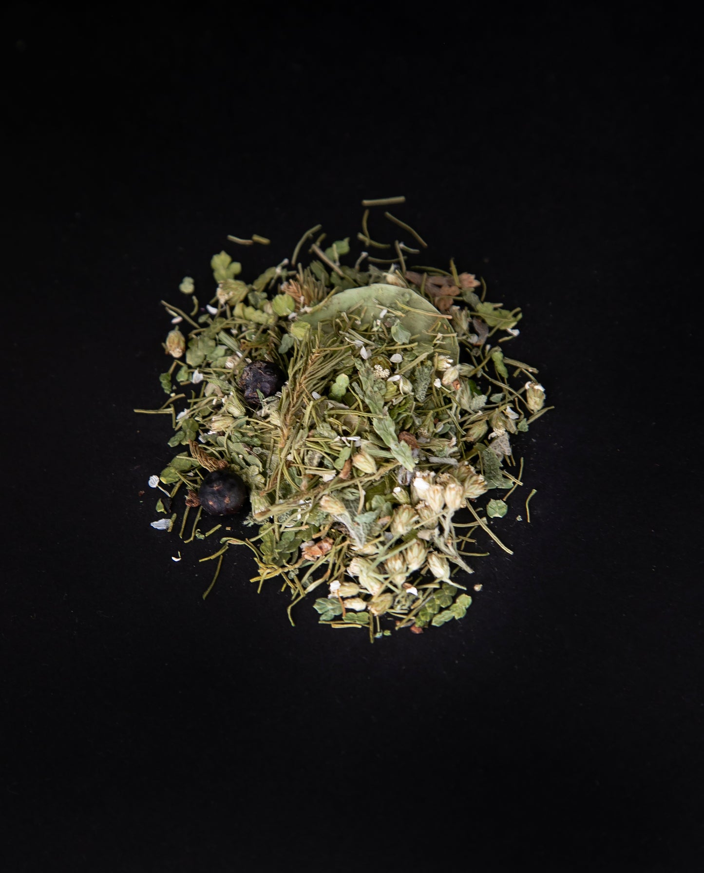 Handful of "Vert Forêt" loose leaf tea on a black background. This forest blend is green and includes visible pieces of spruce tip and juniper berries.