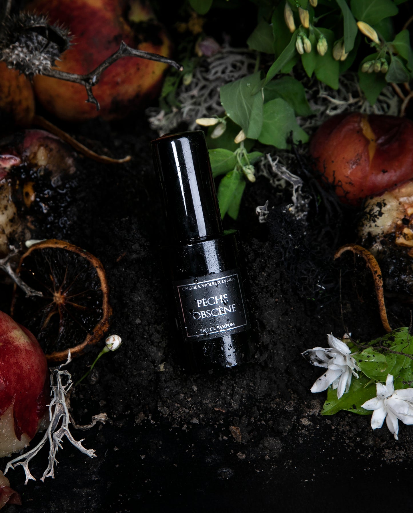 30ml black glass bottle of LVNEA best-selling natural perfume Peche Obscene lying on dirt surrounded by ripe peaches, jasmine, and botanicals