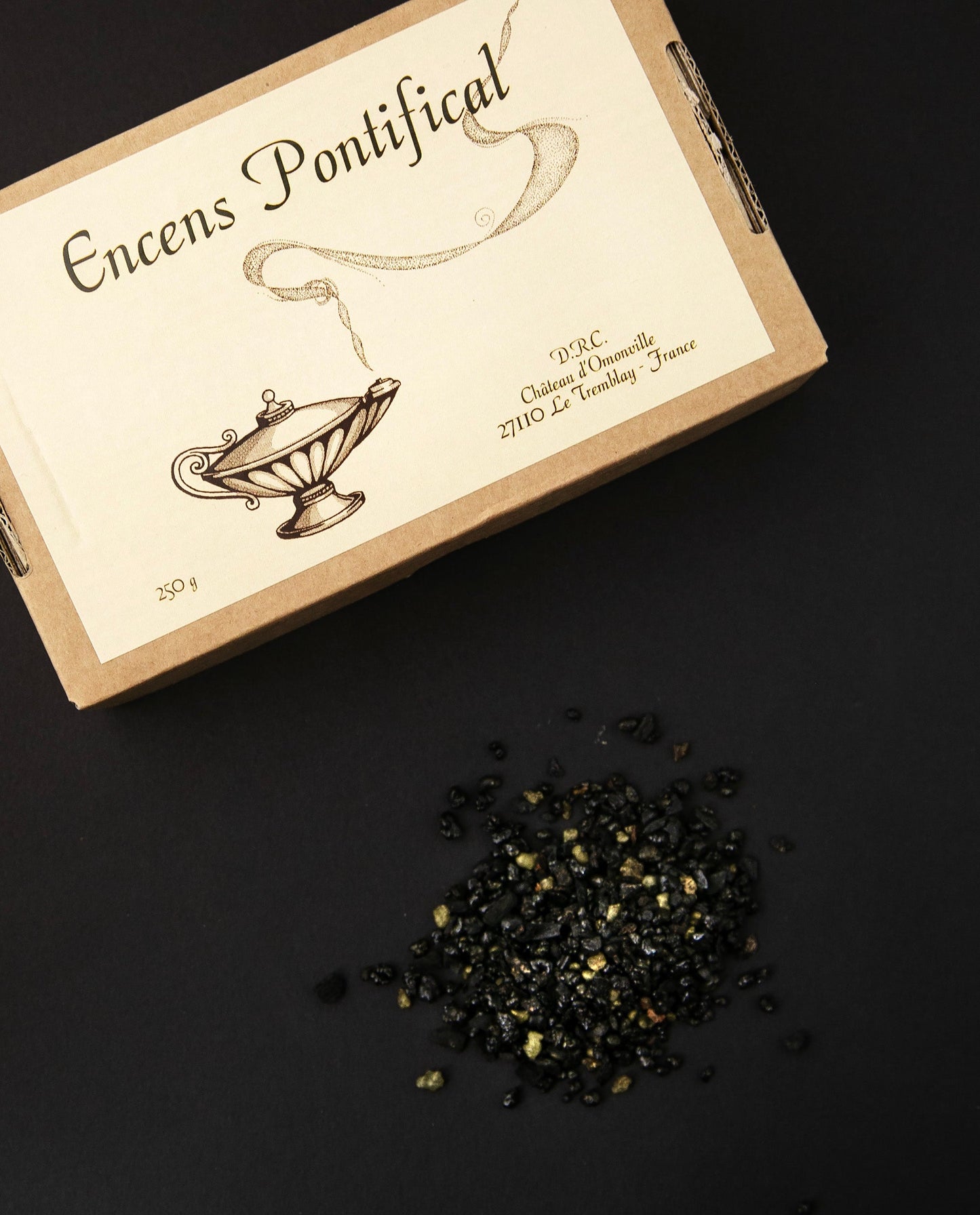 Brown cardboard box of Pontifical Incense. There is a handful of the loose resin incense on the surface, next to the box